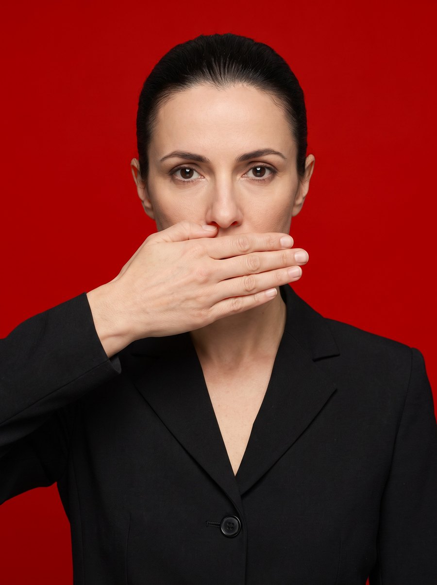 A woman in a black jacket against a deep red ground, her hand flat over her mouth, entirely composed.